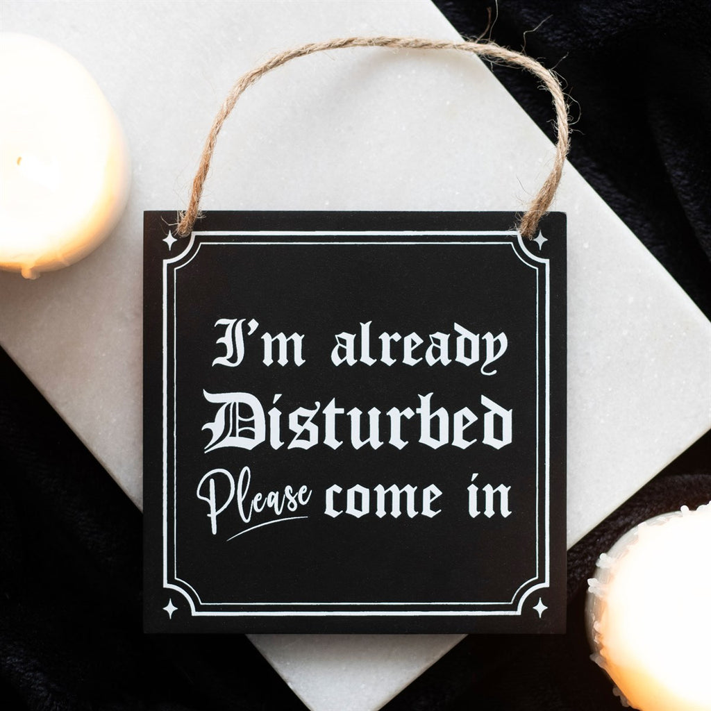 Black wooden hanging sign with white gothic lettering reading I'm Already Disturbed Please Come In, with a jute rope hanger, styled with candles