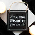 Black wooden hanging sign with white gothic lettering reading I'm Already Disturbed Please Come In, with a jute rope hanger, styled with candles