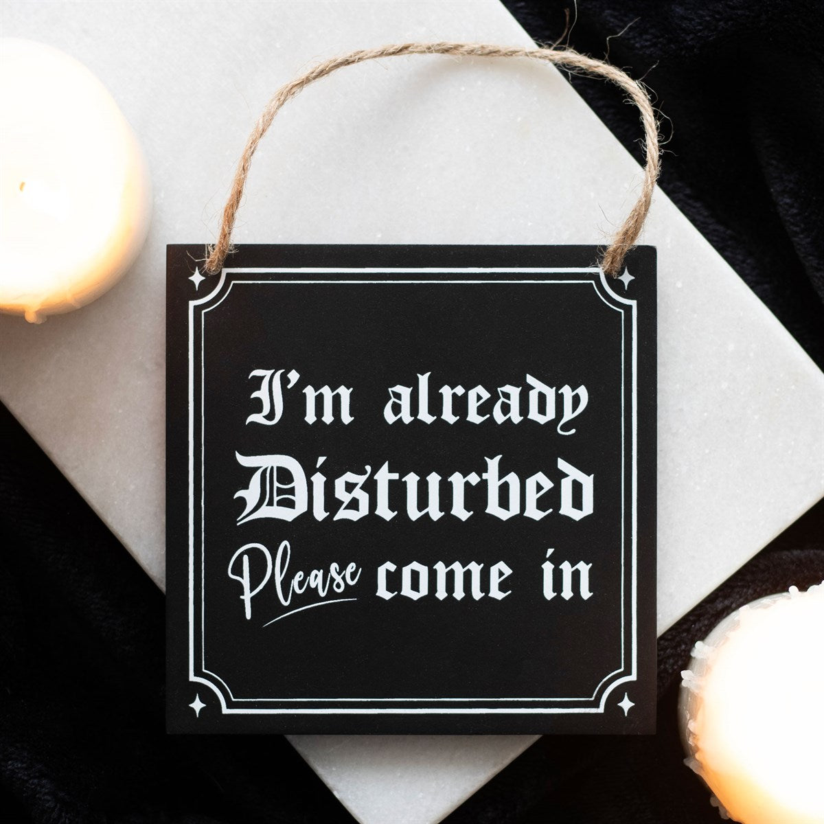 Black wooden hanging sign with white gothic lettering reading I'm Already Disturbed Please Come In, with a jute rope hanger, styled with candles