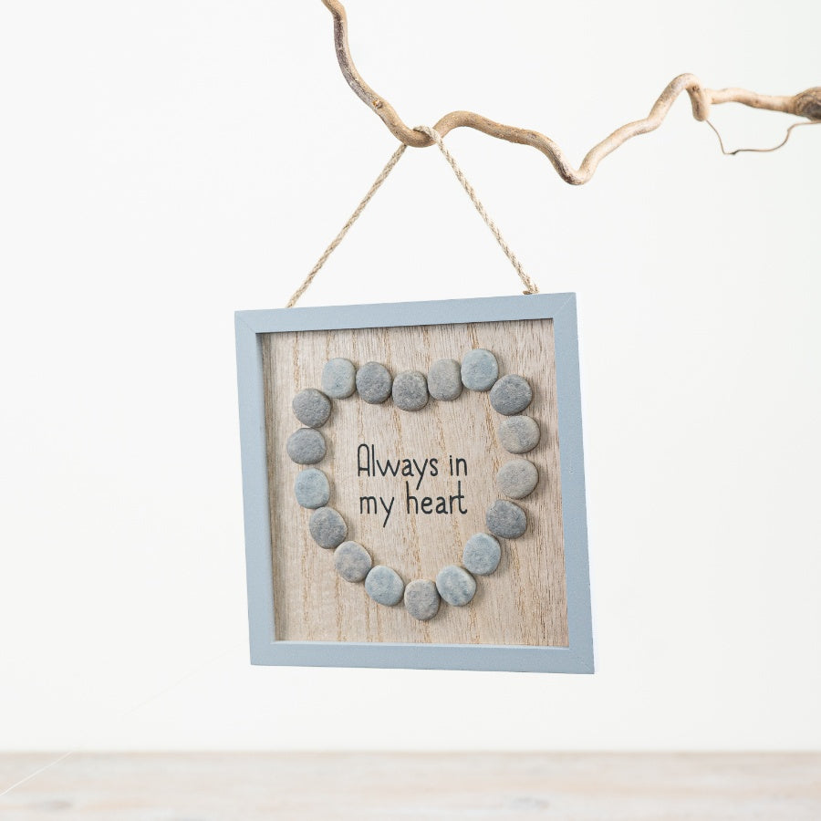 Square hanging wooden frame featuring a heart shape made from smooth grey pebbles surrounding the words “Always In My Heart” on a rustic wood-effect background.