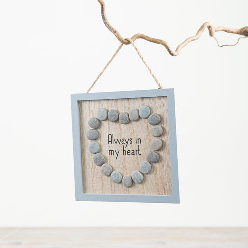 Square hanging wooden frame featuring a heart shape made from smooth grey pebbles surrounding the words “Always In My Heart” on a rustic wood-effect background.