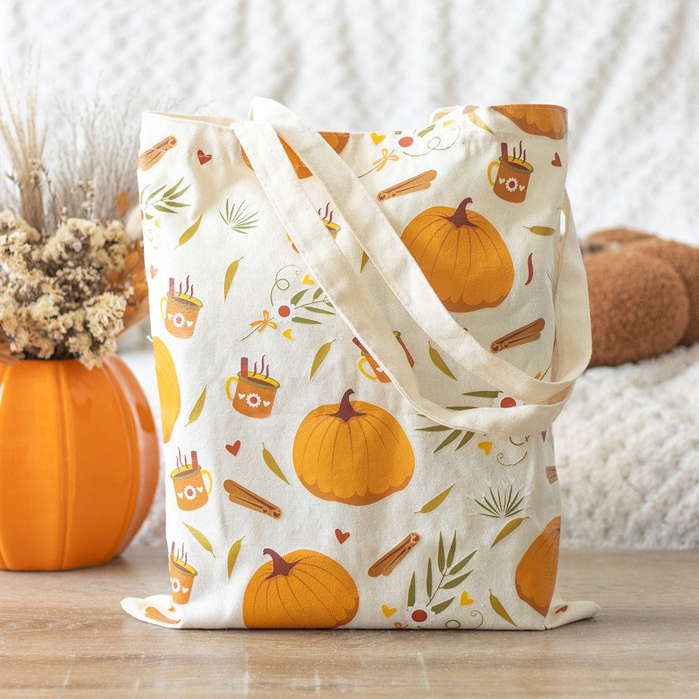 Cream polycotton tote bag with all-over autumn print featuring orange pumpkins hot drink mugs cinnamon sticks and green leaves, styled beside an orange pumpkin vase with dried flowers