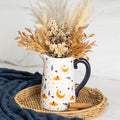 White ceramic flower jug with harvest moon moth and autumn leaves print and navy blue handle, filled with dried pampas grass and autumn foliage on a wicker tray with cinnamon sticks