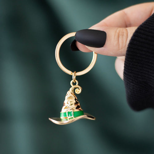 Gold-tone metal witch's hat keyring with green enamel band detail on a gold split ring, held against a dark green background