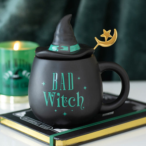 Black matte ceramic Bad Witch mug with teal green lettering, witch hat shaped lid with green band and gold moon and star spoon, styled on a book with a green candle in the background