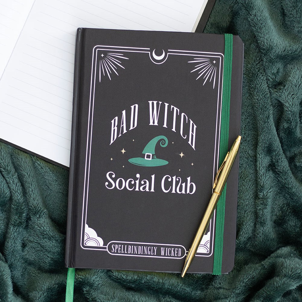 Dark hardback A5 notebook with Bad Witch Social Club design, teal green witch hat illustration and Spellbindingly Wicked text, styled on a green fluffy blanket with a gold pen