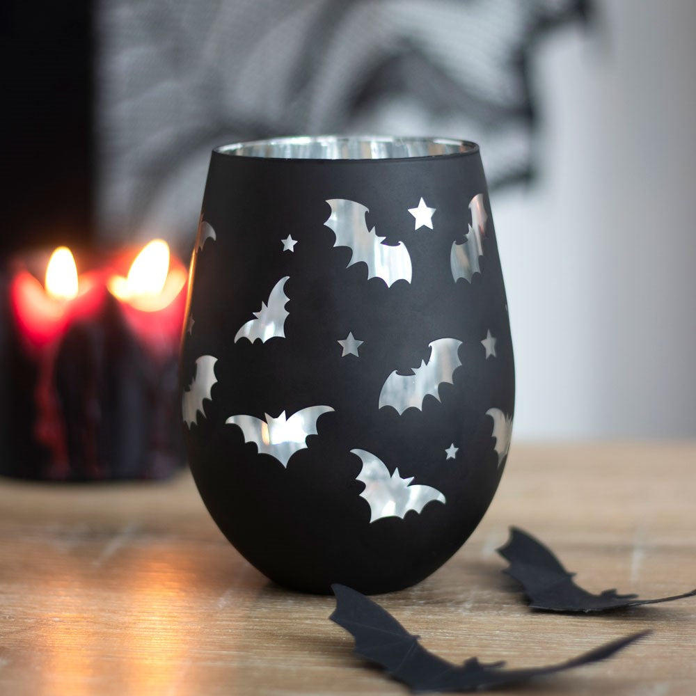 Black stemless wine glass with bat and star cut-out pattern revealing a silver interior, styled on a wooden surface with red candles in the background