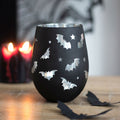 Black stemless wine glass with bat and star cut-out pattern revealing a silver interior, styled on a wooden surface with red candles in the background