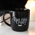 Black ceramic mug featuring “Bat Shit Crazy” text with gothic bat-themed design and classic mug shape.
