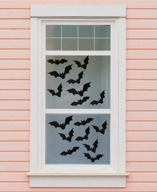 24 black bat window cling decorations in four different sizes applied to a house window, shown from outside against a pink painted wall