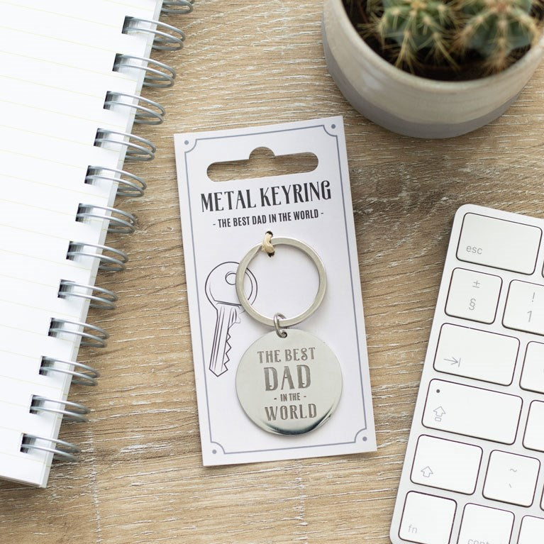Metal keyring with 'The Best Dad in the World' text on a card, placed on a wooden surface with a keyboard and cactus in the background.
