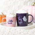 Pink Little Boo and purple Big Boo ceramic Halloween mugs filled with whipped cream, displayed on a white tray with orange and pink pumpkins in the background