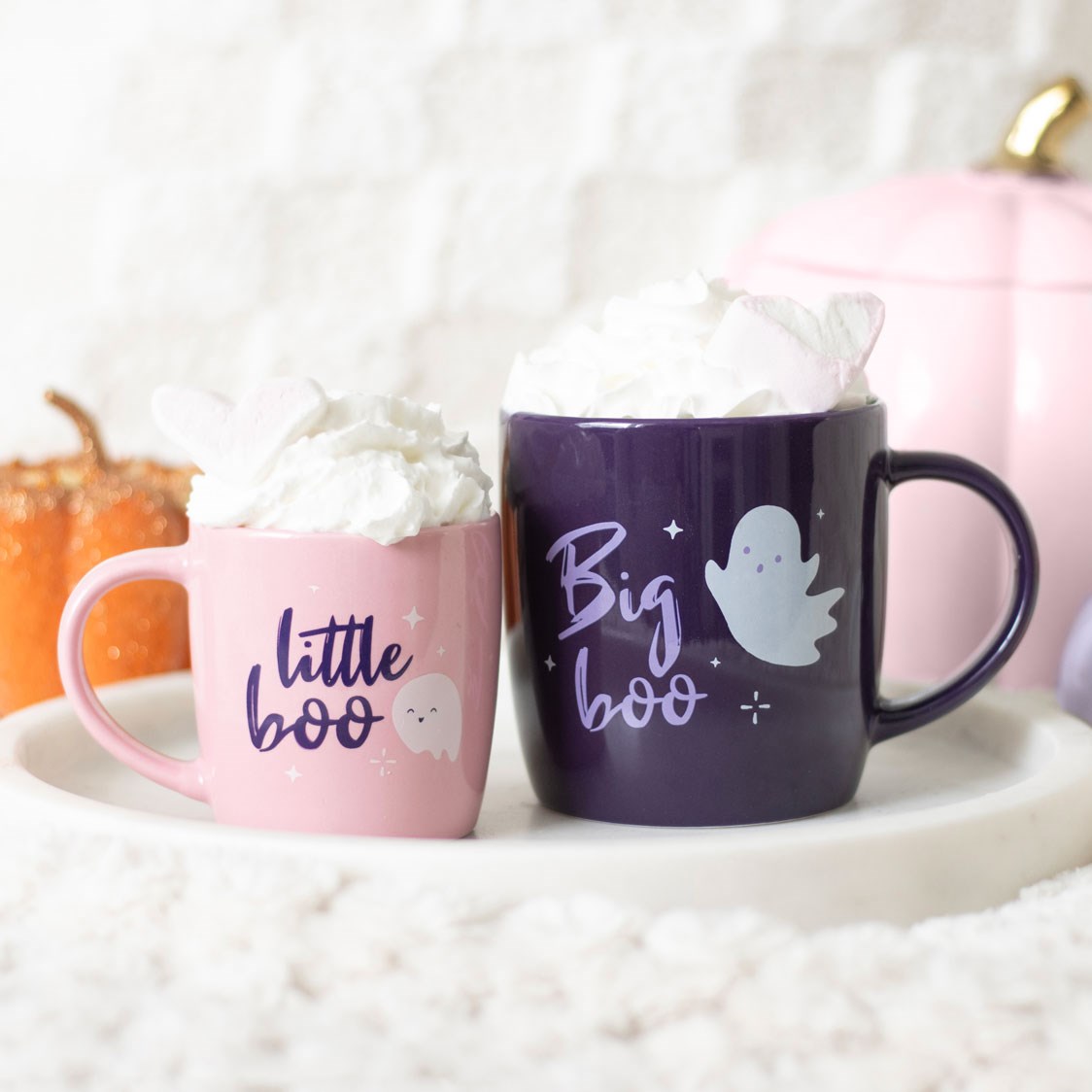 Pink Little Boo and purple Big Boo ceramic Halloween mugs filled with whipped cream, displayed on a white tray with orange and pink pumpkins in the background