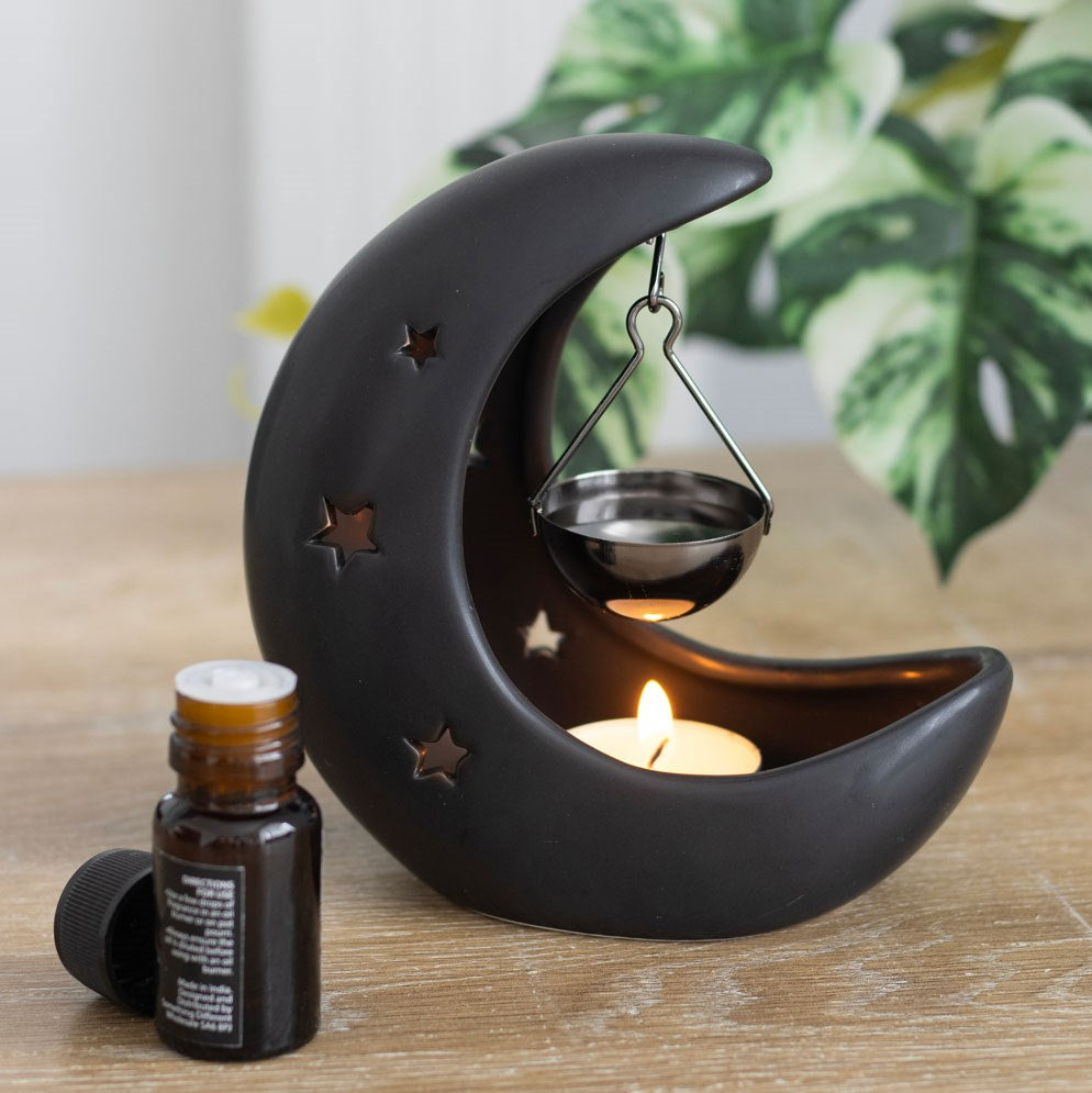 Black crescent moon ceramic oil burner with star cut-outs, hanging metal dish, lit tealight and essential oil bottle on wooden surface
