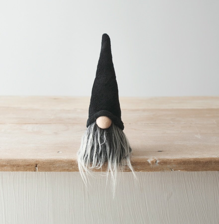 Small black Halloween gonk with tall pointed black hat and long grey beard, sitting on the edge of a rustic wooden shelf against a light grey background