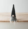 Small black Halloween gonk with tall pointed black hat and long grey beard, sitting on the edge of a rustic wooden shelf against a light grey background