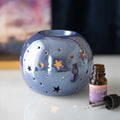 Iridescent blue-grey ceramic round oil burner with star cut-outs, warm candlelight glow, styled with an essential oil bottle on a white surface