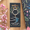 Blue enamel swallow charm keyring on a silver ring, displayed on a black folk art gift card reading "Spread Your Wings Enamel Keyring", laid on a wooden tray with pink gypsophila flowers