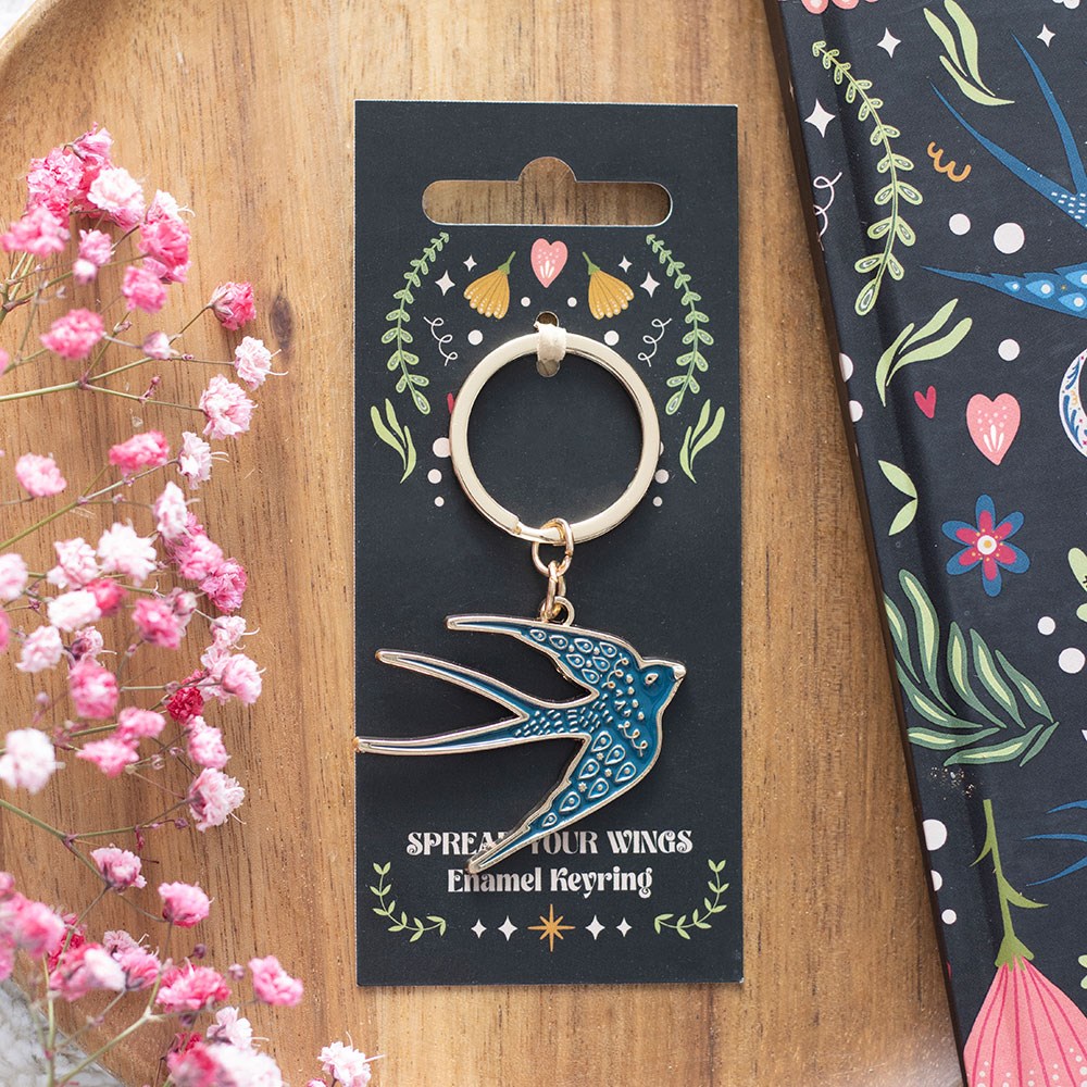 Blue enamel swallow charm keyring on a silver ring, displayed on a black folk art gift card reading "Spread Your Wings Enamel Keyring", laid on a wooden tray with pink gypsophila flowers