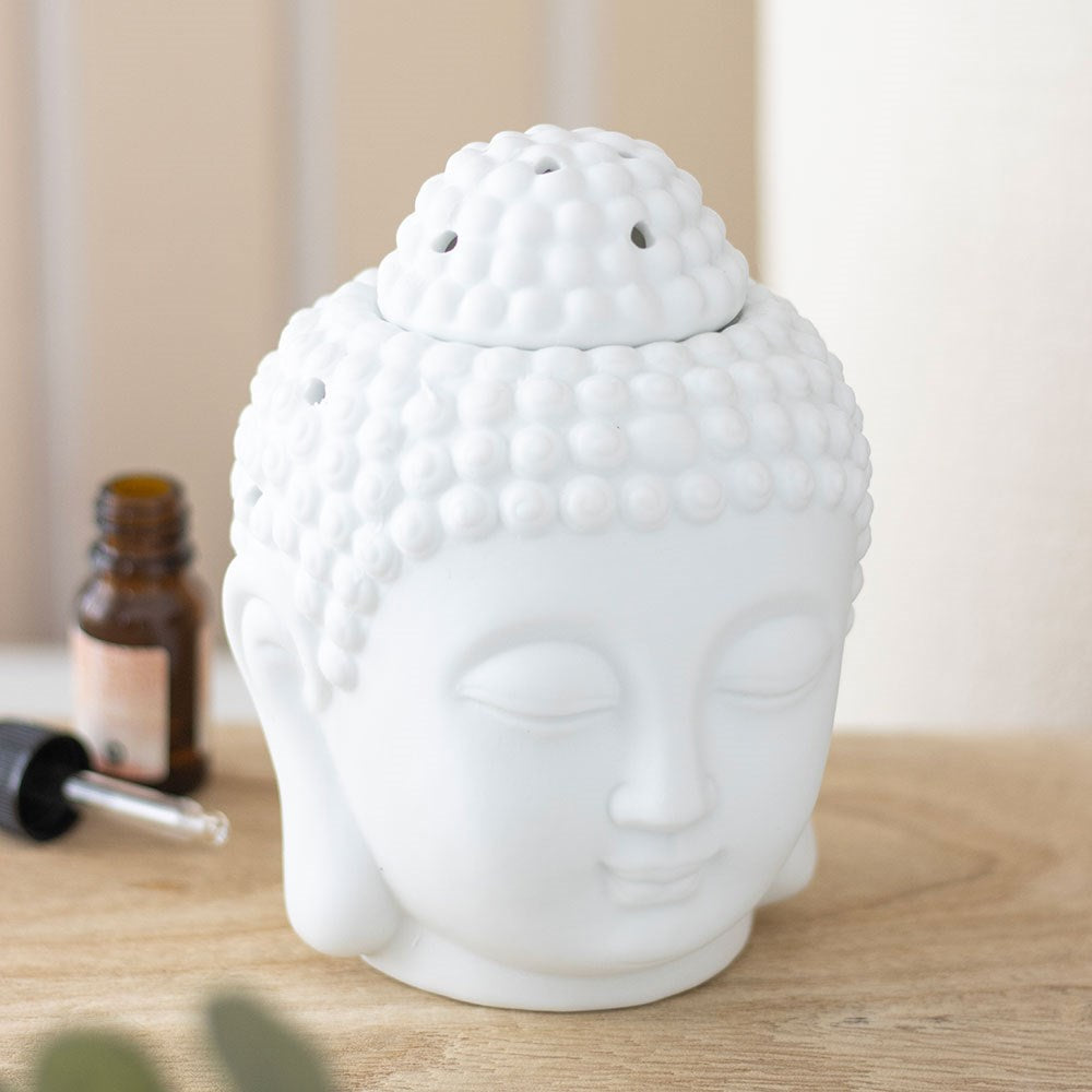 White matte ceramic Buddha head oil burner with removable dish top, styled on a wooden surface with an essential oil bottle