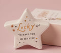 White ceramic star-shaped token with gold Lucky script and floral details on a pink background, with matchbox gift packaging behind