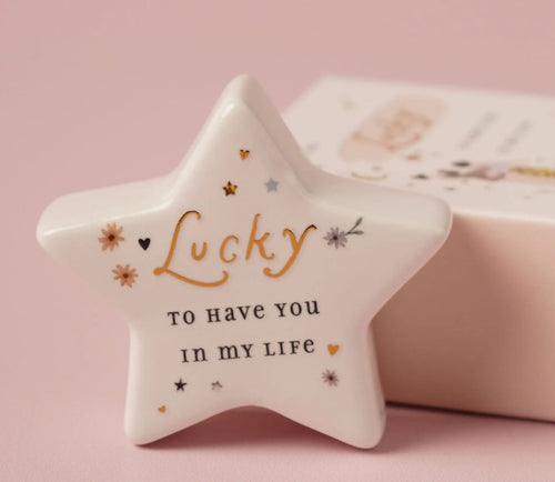 White ceramic star-shaped token with gold Lucky script and floral details on a pink background, with matchbox gift packaging behind