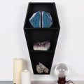 Black wooden coffin-shaped wall shelf with three tiers displaying blue agate bookends, amethyst cluster and sage bundles, styled with white candles and a crystal ball