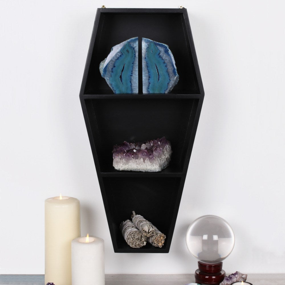 Black wooden coffin-shaped wall shelf with three tiers displaying blue agate bookends, amethyst cluster and sage bundles, styled with white candles and a crystal ball
