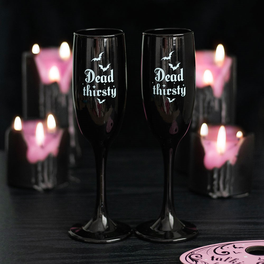 Pair of black gothic champagne flutes with Dead Thirsty text and bat illustrations, styled on a dark surface with pink candles in the background
