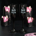 Pair of black gothic champagne flutes with Dead Thirsty text and bat illustrations, styled on a dark surface with pink candles in the background