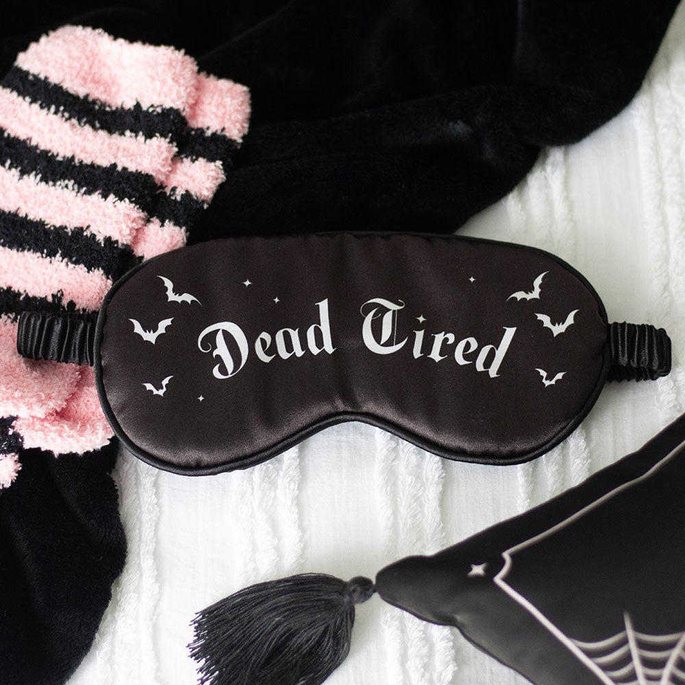 Black satin sleep mask with Dead Tired gothic lettering and bat illustrations, styled on a white knit blanket with pink striped socks