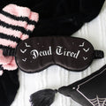 Black satin sleep mask with Dead Tired gothic lettering and bat illustrations, styled on a white knit blanket with pink striped socks
