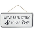 White rectangular metal hanging sign with the phrase "We've Been Dying To See You" and a witch flying on a broomstick illustration.