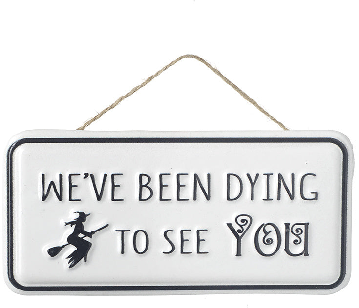 White rectangular metal hanging sign with the phrase "We've Been Dying To See You" and a witch flying on a broomstick illustration.
