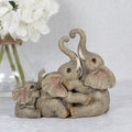 Resin elephant family ornament showing three elephants with raised and entwined trunks in a playful embrace, in natural grey tones, styled with white hydrangeas in the background