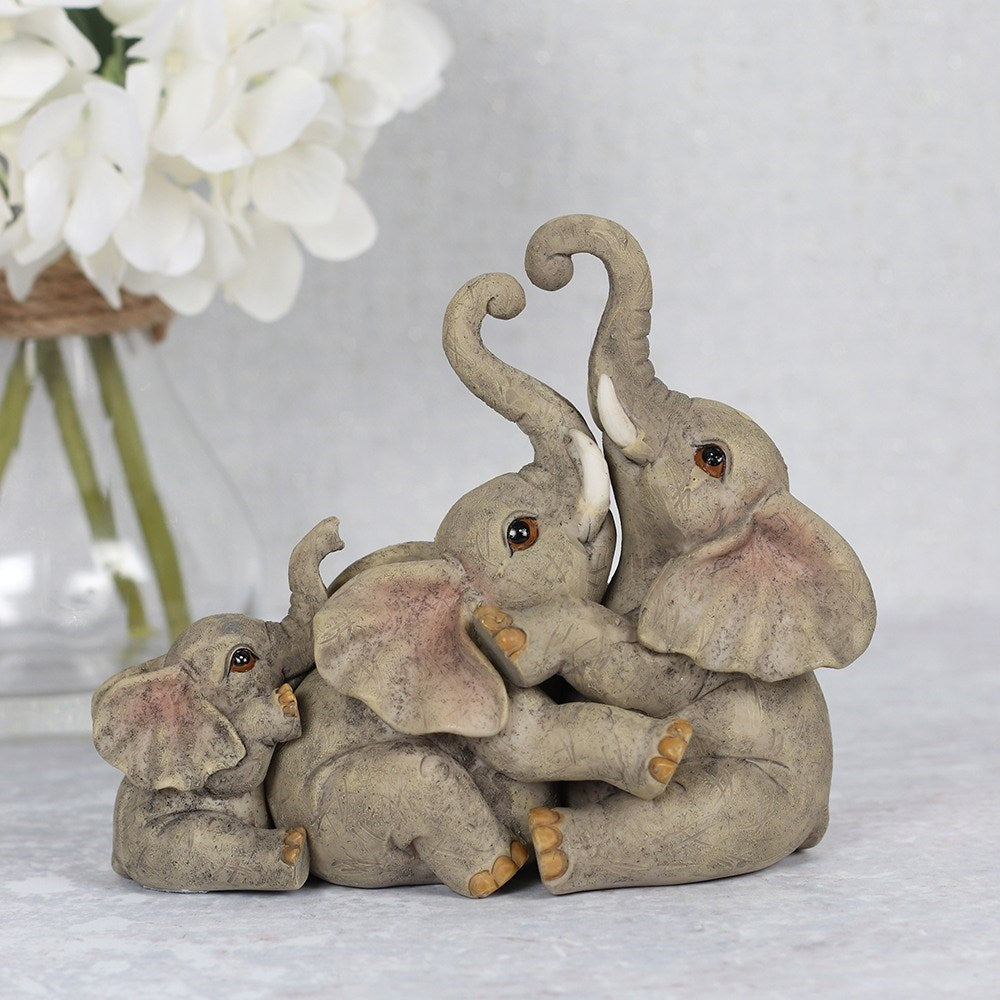 Resin elephant family ornament showing three elephants with raised and entwined trunks in a playful embrace, in natural grey tones, styled with white hydrangeas in the background