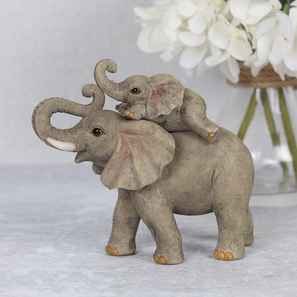 Resin elephant ornament showing a baby elephant perched on its parent's back with both trunks raised, in natural grey tones with a rustic textured finish, styled with white hydrangeas in the background