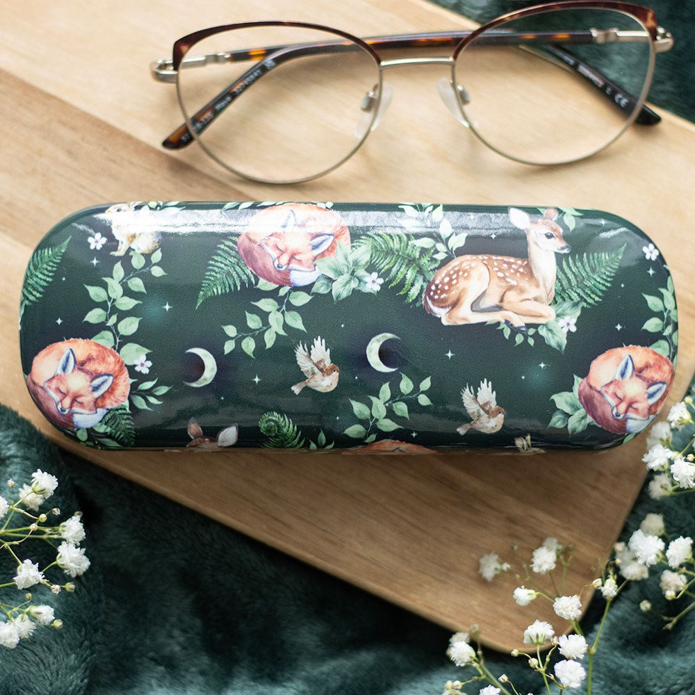 Dark green hard shell glasses case with a woodland pattern featuring sleeping foxes, a resting fawn, birds, fern leaves and crescent moon motifs, displayed on a wooden surface with a pair of glasses resting on top and white gypsophila flowers in the foreground