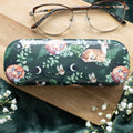 Dark green hard shell glasses case with a woodland pattern featuring sleeping foxes, a resting fawn, birds, fern leaves and crescent moon motifs, displayed on a wooden surface with a pair of glasses resting on top and white gypsophila flowers in the foreground