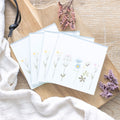 Set of four square glass coasters decorated with delicate wildflower illustrations in soft pastel colours, displayed on a wooden board with dried flowers.