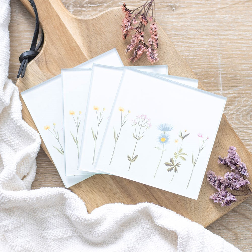 Set of four square glass coasters decorated with delicate wildflower illustrations in soft pastel colours, displayed on a wooden board with dried flowers.