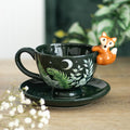 Dark green ceramic teacup and saucer with fern leaf moon and star design and a 3D orange fox figurine perched on the rim, displayed on a wooden surface with white gypsophila flowers