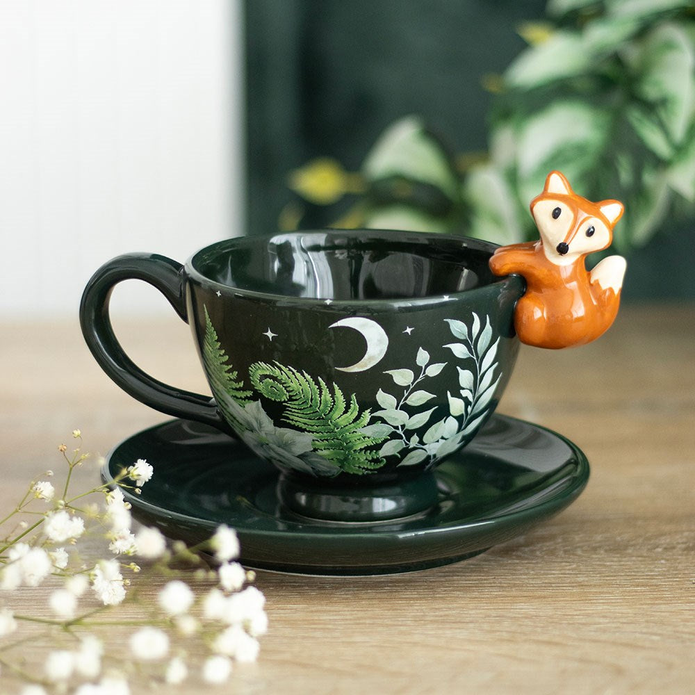 Dark green ceramic teacup and saucer with fern leaf moon and star design and a 3D orange fox figurine perched on the rim, displayed on a wooden surface with white gypsophila flowers
