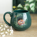 Dark green ceramic rounded mug with a sleeping fox curled in a botanical leaf wreath design, displayed on a wooden surface with white gypsophila flowers and green plants in the background