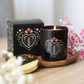 Black glass scented candle with a folk art floral heart design and gold lid, displayed lit on a wooden board next to its matching black gift box, with pink gypsophila flowers in the background