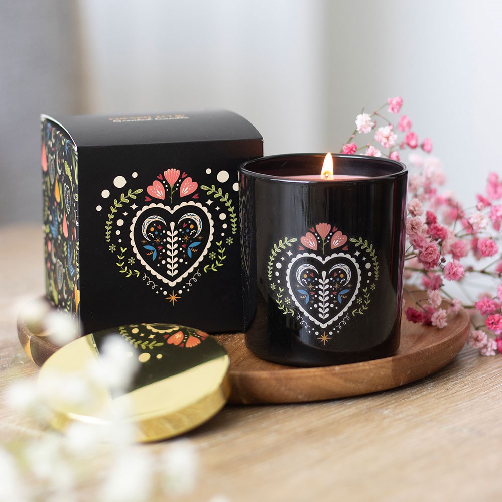 Black glass scented candle with a folk art floral heart design and gold lid, displayed lit on a wooden board next to its matching black gift box, with pink gypsophila flowers in the background