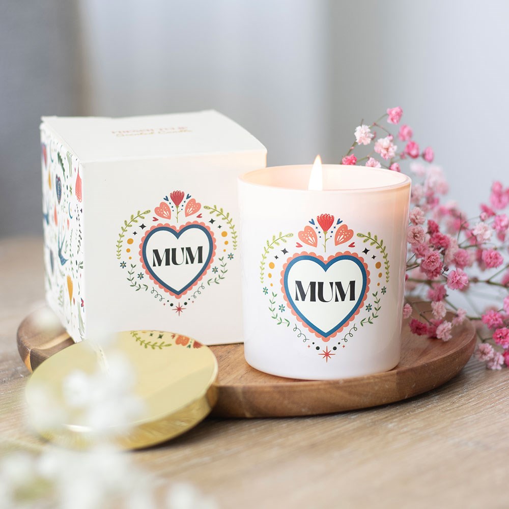 White glass scented candle with a colourful folk art floral heart design reading "Mum", displayed lit on a wooden board next to its matching white gift box, with pink gypsophila flowers