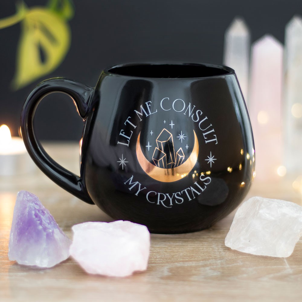 Black rounded ceramic mug featuring the phrase “Let Me Consult My Crystals” with a crescent moon, crystal illustration and star details.