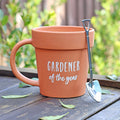 Ceramic plant pot shaped ceramic mug with "Gardener of the Year" text in white, displayed outdoors on a wooden surface with a stainless steel shovel spoon leaning against it and scattered leaves