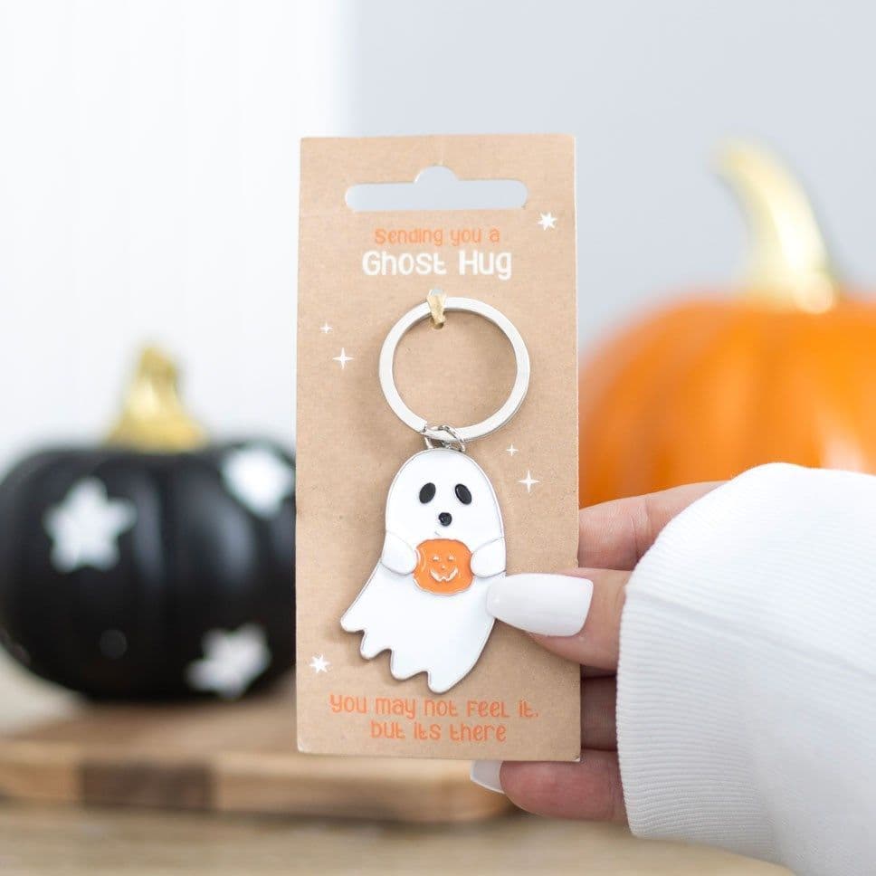 White ghost metal and enamel keyring holding an orange jack-o-lantern pumpkin, displayed on a kraft Sending You A Ghost Hug sentiment card, held against a Halloween pumpkin backdrop